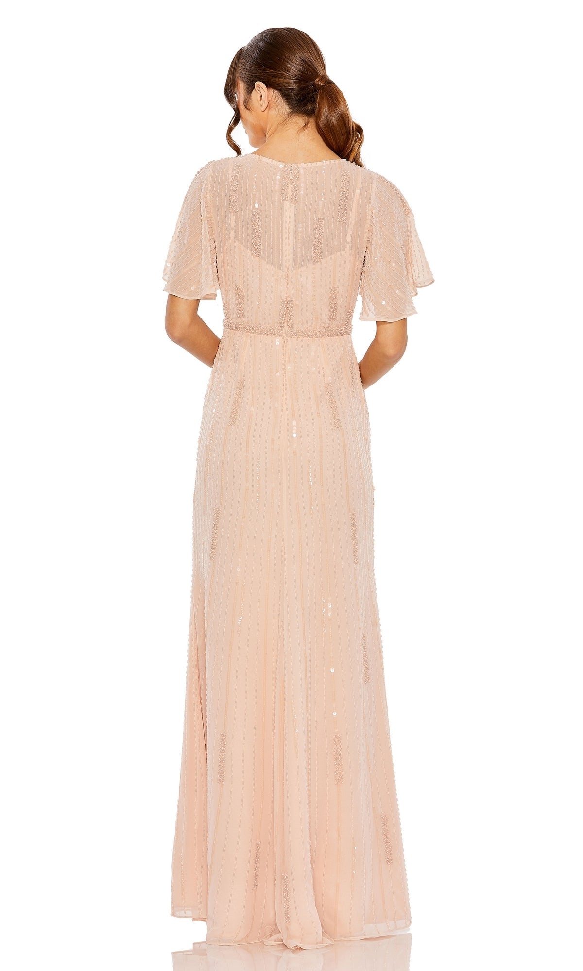 A woman faces away, wearing the Mac Duggal Long Formal Dress 11014—a blush pink, floor-length gown with a v-neckline, sheer short sleeves, sequin accents, and a sheer overlay. Her hair is styled in a low ponytail.